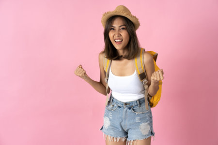 Young woman going on vacation wearing hat and backpack and clenching fists in excitement against...
