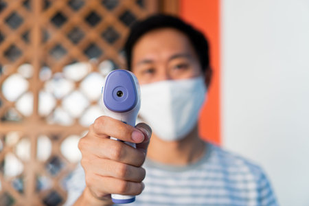 Young man in face mask using medical digital infrared thermometer to check and monitor body...