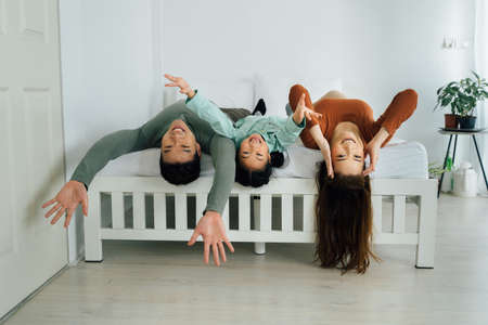 Cheerful Asian mother and father with young daughter lying on backs on bed and looking at camera. Family togetherness and bonding indoors conceptの写真素材