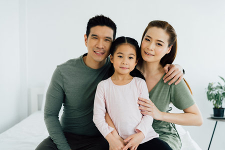 Smiling happy Asian mother and father looking at camera with young daughter in white room...
