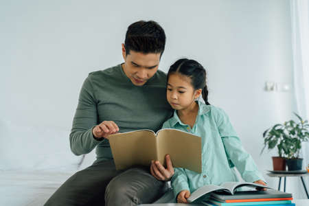 Authentic Asian father sitting with young daughter looking at book in the room background. Homeschooling concept.の写真素材