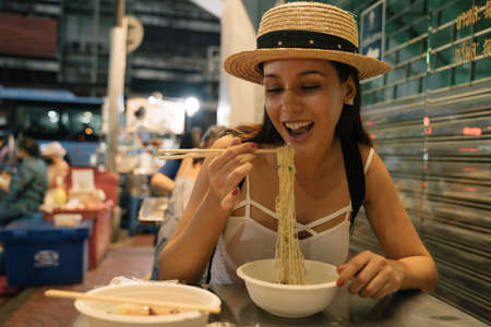 Cheerful young Hispanic Brunette woman sitting in noodle bar using chopsticks to eat bowel of freshly made noodlesの写真素材