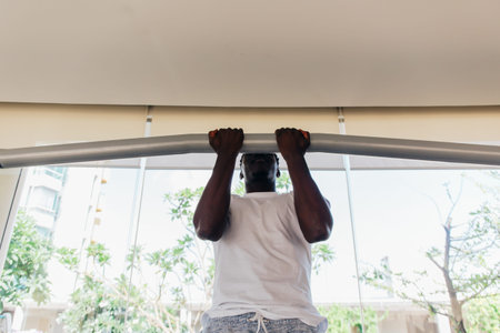 Strong African American sportsman in white T-shirt doing pull ups during fitness workout in modern...