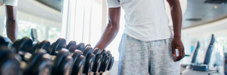 African American man picking up a dumbbell from the rack and looking outside at gym. Male weight training person holding sport equipment with serous look in fitness centerの写真素材