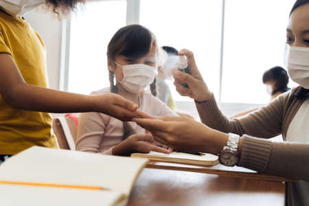 Young female teacher using an alcohol spray to disinfect student hands in classroom. Asian woman in face mask cleaning pupils hands with hand sanitizer. School reopen after quarantine and lockdown.の写真素材