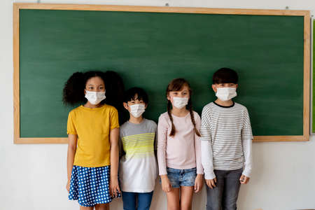 Group of diverse elementary students in classroom. Primary school multi-racial pupils standing in a row wearing a face mask with blackboard in background. School new normal conceptの写真素材