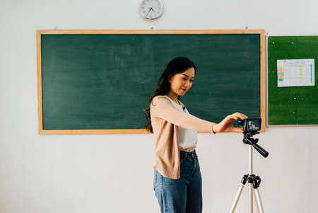 Young woman setting up video camera for online tutoring, technology, remote learning, online education. Asian school teacher preparing to deliver online courseの写真素材