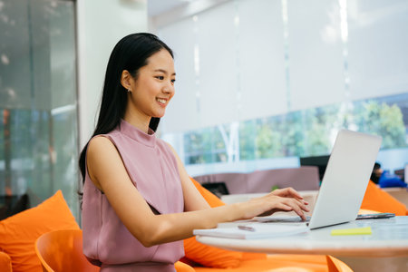 Cheerful Asian businesswoman working on laptop at corporate workplace. Happy and successful white...