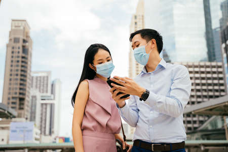 Asian businessman and woman wearing Covid face coverings looking at cell phone in urban setting, city life, pandemic, working life, colleagueの写真素材