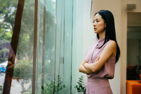Asian businesswoman with arms folded looking through large window at work, contemplation, aspiration, ideasの写真素材