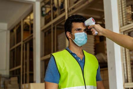 Young Indian factory warehouse worker wearing face mask getting body temperature check with infrared thermometer scanner before working. Security measure at workplace in Coronavirus Covid 19 pandemicの写真素材