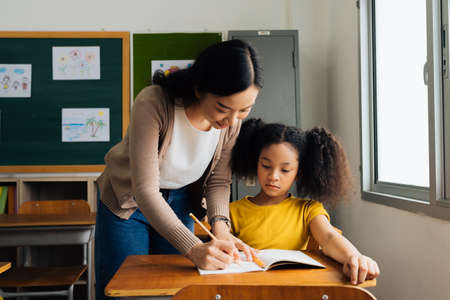 Asian school teacher assisting female student in classroom. Young woman working in school helping girl with her writing, education, support, care.の写真素材