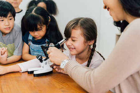 Young woman showing multiethnic children microscope in science class, learning, discovery, curiosity. Asian school teacher helping students use microscopeの写真素材