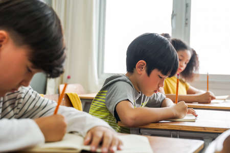 Asian school children sitting at desk in school writing in note book with pencil, studying, education, learning. Male and female students in classroom. Boy and girl in elementary ageの写真素材
