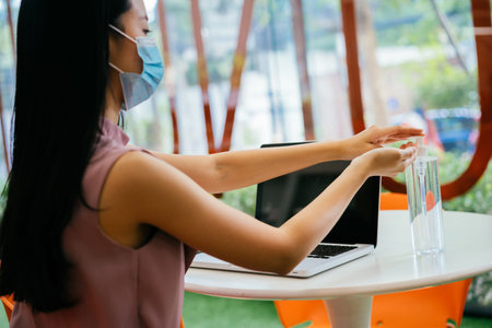Asian businesswoman wearing protective Covid face covering working in modern office, using computer,...