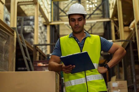 Young Indian industrial factory warehouse worker working in logistic industry indoor. Thoughtful serious man holding a clipboard checking item merchandise stock order in storehouseの写真素材