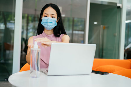 Asian businesswoman wearing protective Covid face covering working in modern office, using computer,...