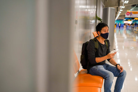 20s young adult Asian male tourist backpacker holding flight ticket wearing a face mask with a flight ticket at the airport. Social distancing travel safety advice during Covid-19 concept