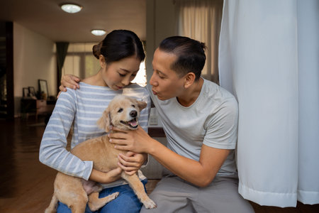 Young adult Asian couple holding a puppy with home interior in background. 30s mature man kissing a dog pet in arms of woman. Husband and wife living together in the house. Modern family happinessの写真素材