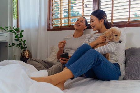 Portrait of happy young adult Asian couple on bed along with dog pet in bedroom interior scene. Man...