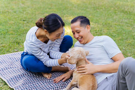Young adult Asian couple playing a puppy with green grass outdoors in background. 30s mature man and woman lying down on mat in the park touching a dog pet. Happy family and modern life conceptの写真素材