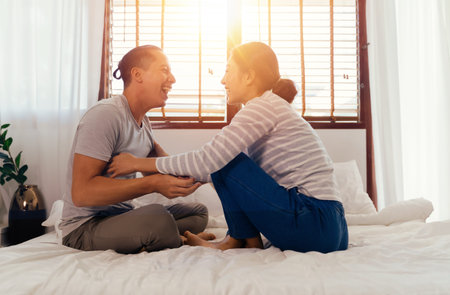 Portrait of happy young adult Asian couple tickling each other on bed together in bedroom interior scene. 30s candid mature husband and wife smiling. Marriage and happy relationship life concept.の写真素材