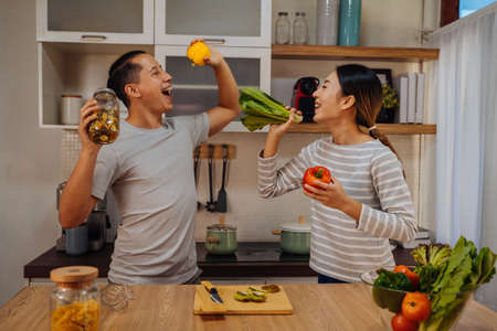 Young lovely couple dancing in the kitchen with window. Asian woman and Caucasian man singing with vegetables and ingredients in modern homeの写真素材