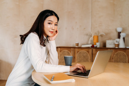 Young teenage Asian woman wears sweater with pretty smile in living room. Happy and successful girl using laptop with notebook.Online technology concept. Winter portrait fashion lifestyle. Copy space.の写真素材