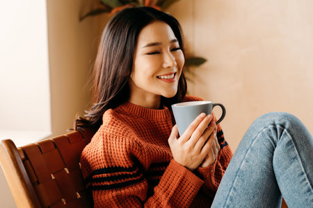 Delighted young ethnic female in sweater smiling and enjoying hot beverage while resting on comfortable chair at homeの写真素材