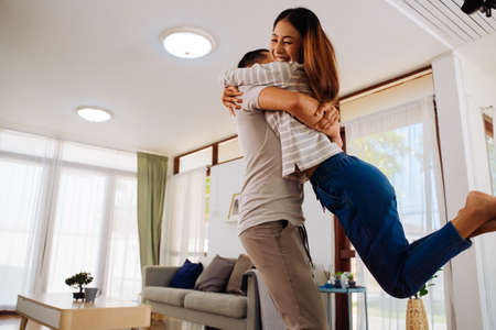 30s young adult Asian man holding woman in the living room at home. Happy couple turning and spinning around with excitement in cozy house. Happy relationship life concept.の写真素材