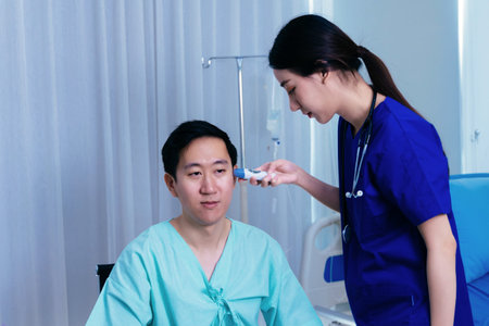 Young female medical worker with stethoscope examining ears of Asian male patient in uniform using...