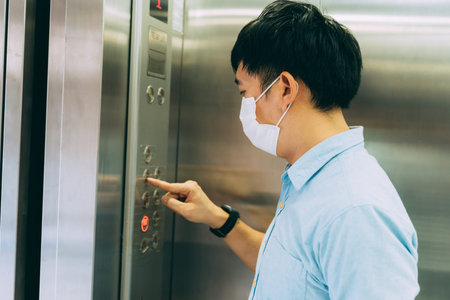 Young Asian businessman wearing protective face mask as precaution against coronavirus standing in elevator pressing floor button to go upstairsの写真素材