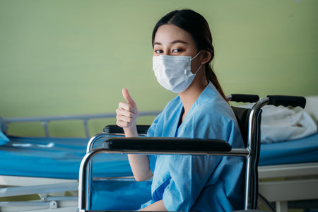 Portrait of a smiling Asian young female patient with surgical mask sitting on wheelchair in...