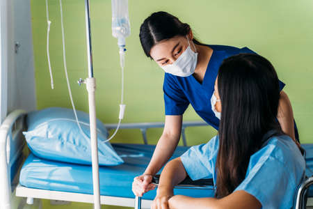 Young nurse wearing surgical mask in conversation with and comforting depressed Asian patient sitting in wheelchair in hospital ward roomの写真素材
