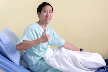 Portrait of a smiling Asian young male patient sitting on hospital bed room showing thumbs up gesture and sign to cameraの写真素材