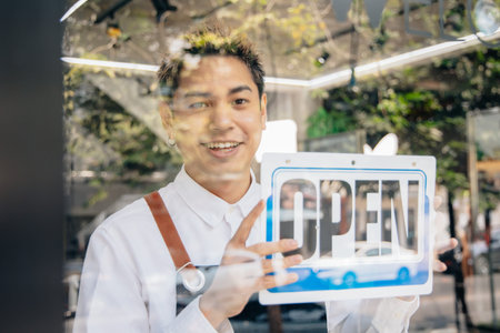 Young Asian man hanging open sign by glass window. Male shop owner holding open notice in front of...