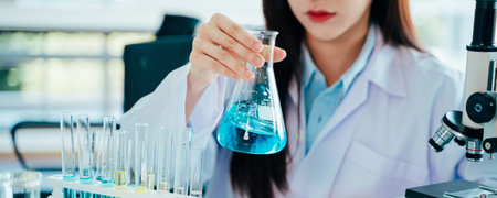 Focused young and beautiful female Asian scientist wearing labcoat and safety eyewear doing medical...