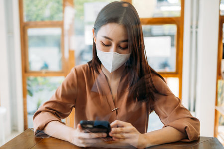 Beautiful young Asian woman sitting in modern cafe wearing face protection mask using and browsing through smartphone while relaxingの写真素材