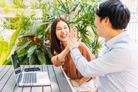 Cheerful and excited young Asian man and woman celebrating good news and success by giving high five to each other while sitting in modern cafeの写真素材