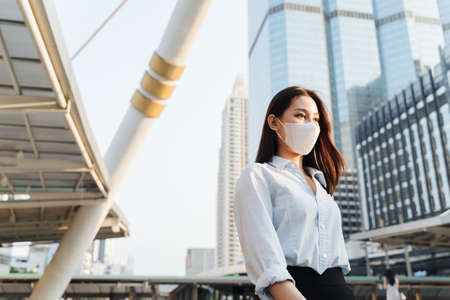 Asian young businesswoman wearing a covid-19 protective mask over her face while walking in streets of office complex at industrial arenaの写真素材