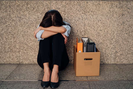 Unrecognized young woman hiding her face after being laid off from job due to economic recession sitting with carton of belongings on floor and cryingの写真素材