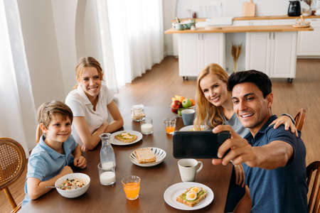Caucasian Family group of four taking selfie photo while eating a breakfast on dining table. Father, Mother, Daughter and Son enjoying happy time together with kitchen in background.の写真素材