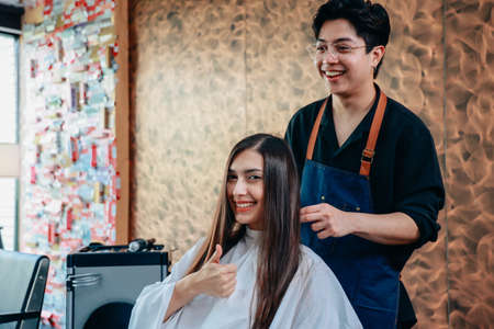 Portrait of happy and cheerful young female customer showing thumbsup sign with male hairdresser wearing apron styling hairの写真素材