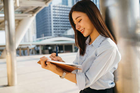 Cheerful young Asian female designer doodling creativity and designs in book while relaxing outside office building during break hoursの写真素材