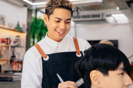 Happy young Asian male hairdresser smiling wearing apron in modern salon holding brush and scissors trimming hair of clientの写真素材