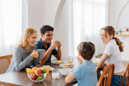 Beautiful and handsome young smiling mother and father sitting on table with daughter and son while the family enjoying healthy breakfast morning at homeの写真素材