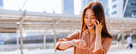 Successful young Asian woman in formal clothing checking time in wristwatch while walking and talking over phone in city street at nightの写真素材