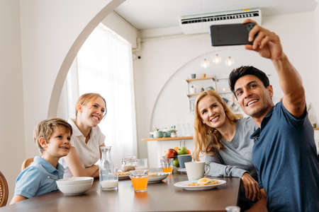 Caucasian Family group of four taking selfie photo while eating a breakfast on dining table. Father, Mother, Daughter and Son enjoying happy time together with kitchen in background.の写真素材