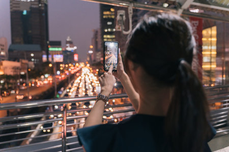 Rear view of young unrecognizable woman standing on footbridge taking picture of evening view of...