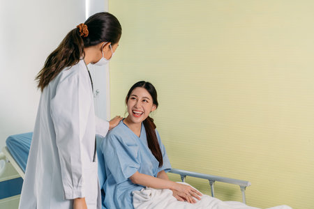 Cheerful young Asian patient sitting on hospital bed laughing and smiling while talking to young female doctor in labcoat wearing face mask for safetyの写真素材